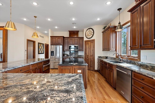 kitchen featuring dark stone counters, a kitchen island with sink, a sink, stainless steel appliances, and light wood-style floors