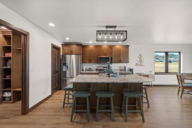 kitchen with a breakfast bar area, an island with sink, light stone counters, light wood-style flooring, and appliances with stainless steel finishes