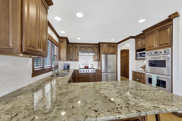 kitchen with a peninsula, light stone countertops, under cabinet range hood, appliances with stainless steel finishes, and a sink