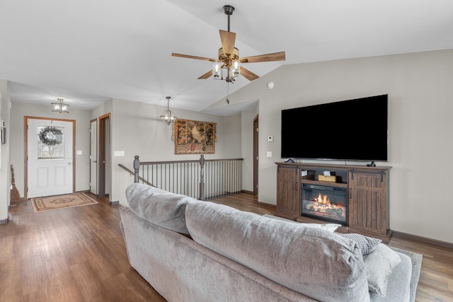 living room with wood finished floors, vaulted ceiling, baseboards, a glass covered fireplace, and ceiling fan with notable chandelier
