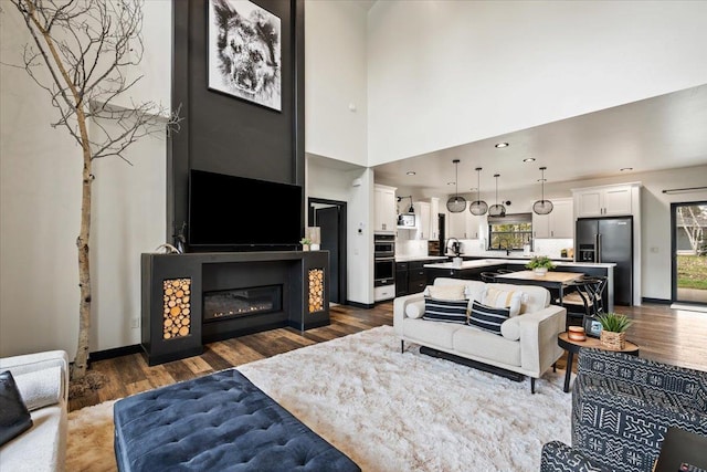 living area featuring healthy amount of natural light, dark wood-style flooring, a glass covered fireplace, recessed lighting, and a high ceiling