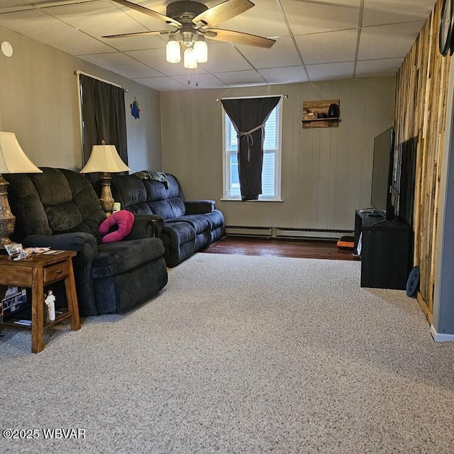 carpeted living area with wood walls, baseboard heating, a paneled ceiling, and a ceiling fan