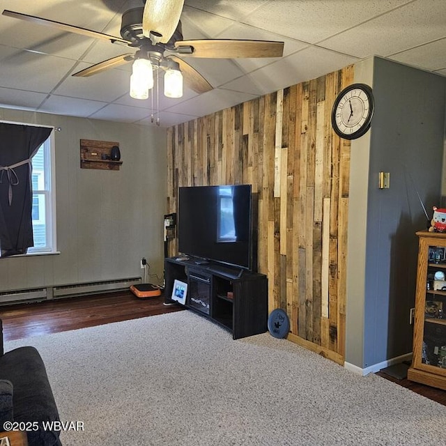 living area with wooden walls, a drop ceiling, a baseboard heating unit, and ceiling fan