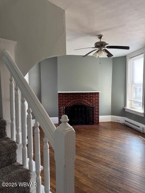 living room featuring a ceiling fan, a baseboard heating unit, wood finished floors, a brick fireplace, and stairs