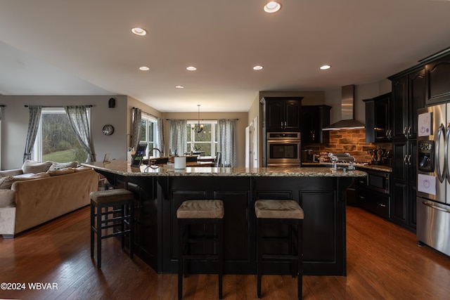 kitchen featuring dark hardwood / wood-style flooring, wall chimney range hood, stainless steel appliances, and a center island with sink