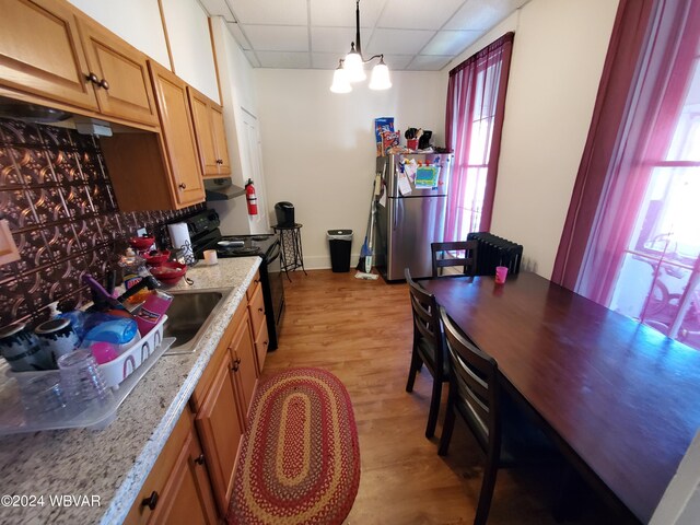 kitchen with a drop ceiling, black electric range oven, hanging light fixtures, light hardwood / wood-style flooring, and stainless steel fridge