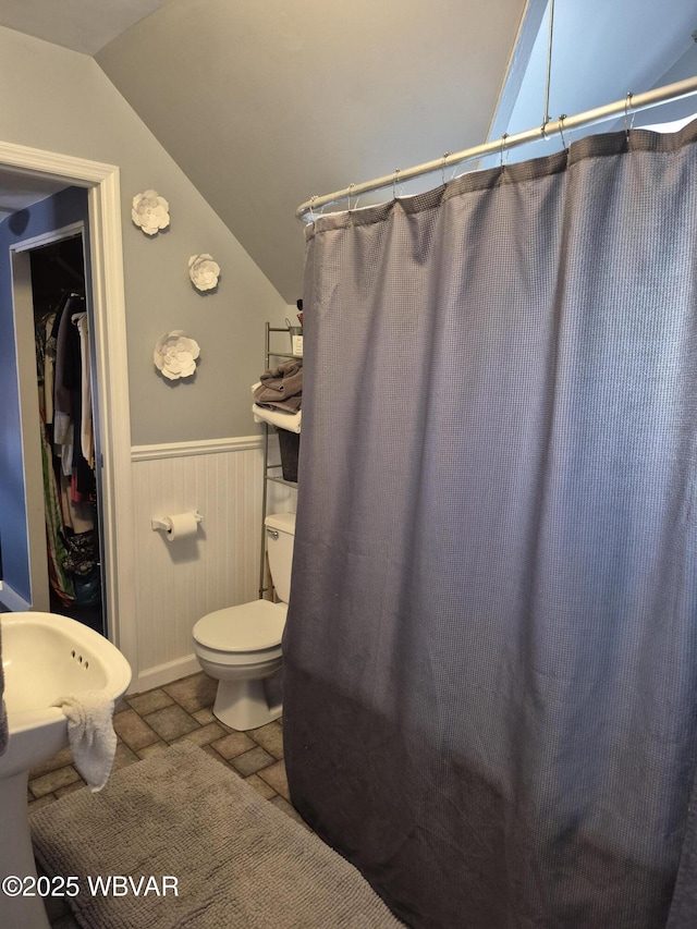 bathroom with lofted ceiling, toilet, stone tile floors, a wainscoted wall, and a shower with curtain