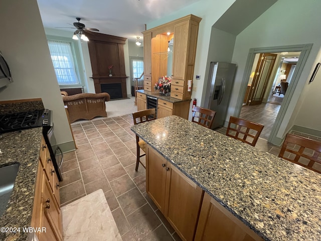 kitchen featuring dark stone counters, stainless steel refrigerator with ice dispenser, ceiling fan, a fireplace, and a breakfast bar area