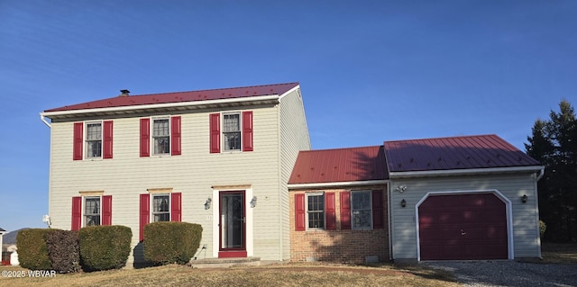 colonial-style house featuring gravel driveway, brick siding, a garage, and metal roof