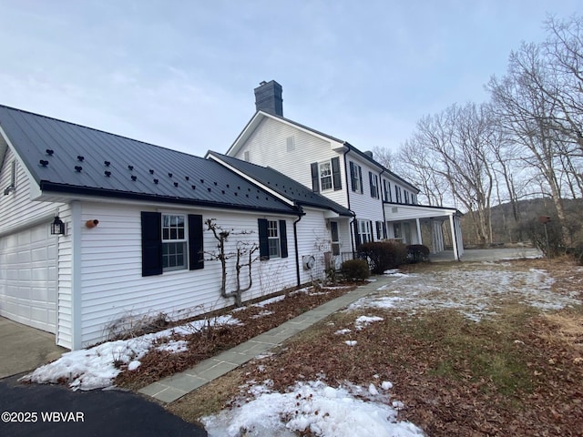 view of snowy exterior featuring an attached garage, a chimney, and metal roof