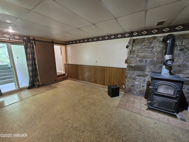 unfurnished living room with a wood stove, wainscoting, a drop ceiling, a barn door, and wooden walls