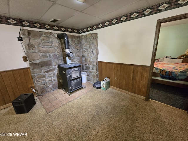 unfurnished living room with a wainscoted wall, a wood stove, a drop ceiling, carpet, and wood walls