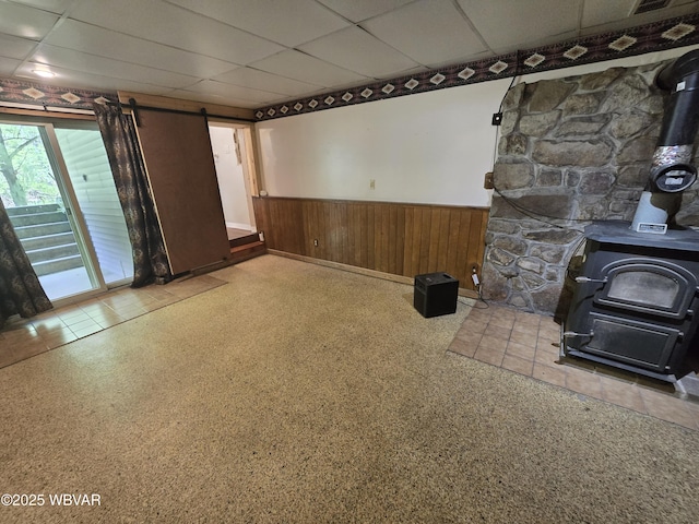 unfurnished living room featuring a wood stove, a wainscoted wall, a paneled ceiling, a barn door, and wood walls