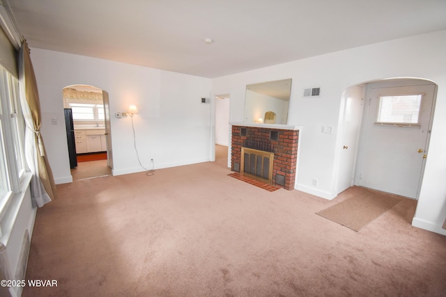 unfurnished living room with visible vents, arched walkways, baseboards, light colored carpet, and a brick fireplace