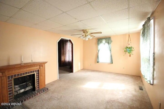 unfurnished living room featuring a drop ceiling, a brick fireplace, ceiling fan, and carpet flooring