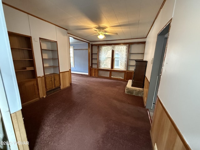 unfurnished living room featuring ceiling fan, wood walls, built in features, and dark colored carpet