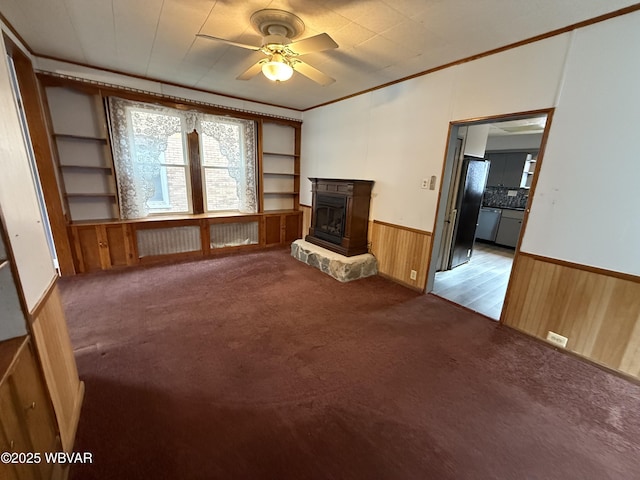 unfurnished living room featuring a stone fireplace, ceiling fan, crown molding, light carpet, and built in shelves