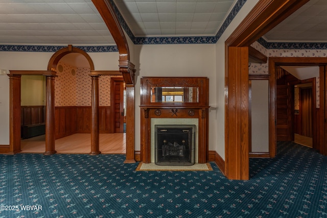 unfurnished living room featuring ornate columns, wooden walls, a tiled fireplace, ornamental molding, and carpet