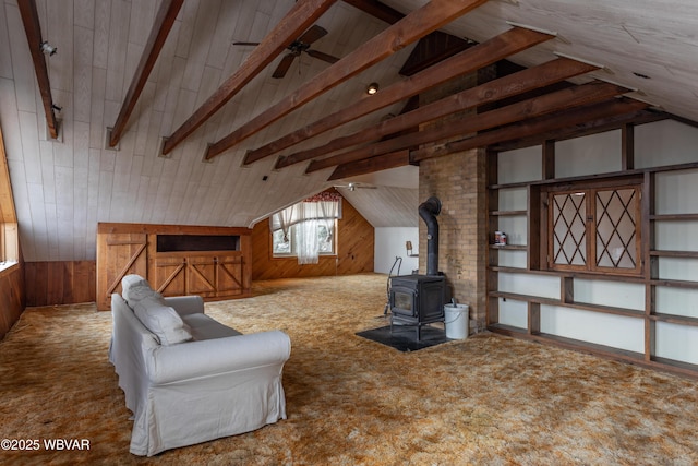 carpeted living room featuring ceiling fan, wooden walls, lofted ceiling with beams, and a wood stove