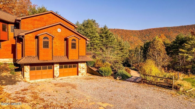 view of front of home with driveway, stone siding, roof with shingles, a wooded view, and an attached garage