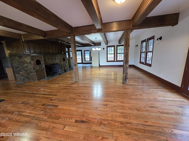 unfurnished living room with beam ceiling, wood finished floors, a stone fireplace, an inviting chandelier, and baseboards