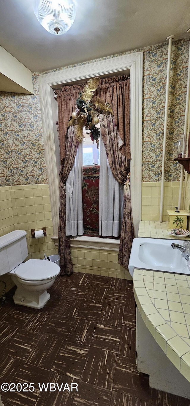 bathroom featuring wallpapered walls, wainscoting, vanity, tile walls, and toilet