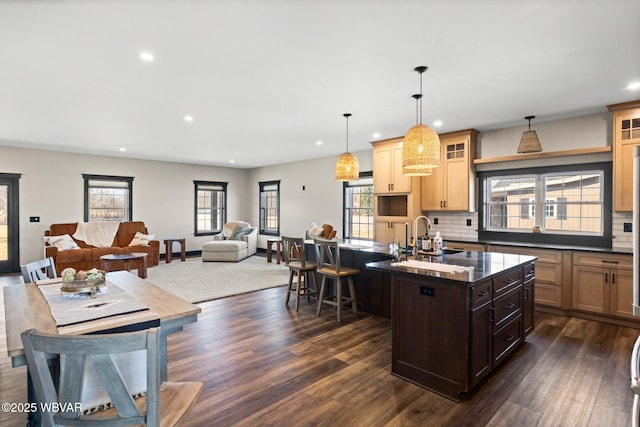 kitchen featuring a kitchen island with sink, a sink, glass insert cabinets, dark wood finished floors, and decorative backsplash