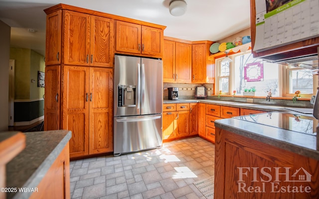 kitchen with backsplash, stone finish floor, brown cabinetry, stainless steel fridge, and black electric cooktop