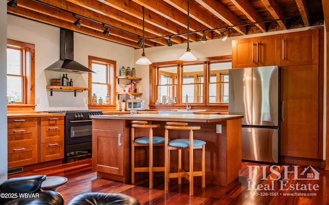 kitchen featuring stainless steel refrigerator, sink, wall chimney range hood, stove, and a kitchen island with sink