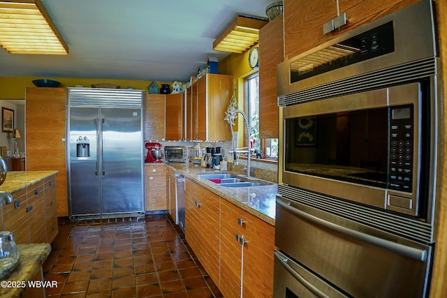 kitchen with a sink, tasteful backsplash, built in appliances, light stone countertops, and brown cabinetry