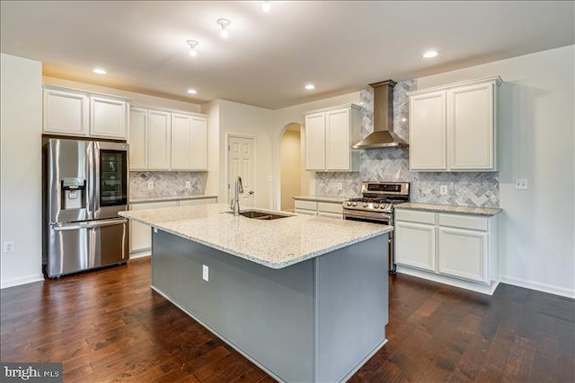 kitchen with dark wood-type flooring, sink, an island with sink, stainless steel appliances, and wall chimney range hood