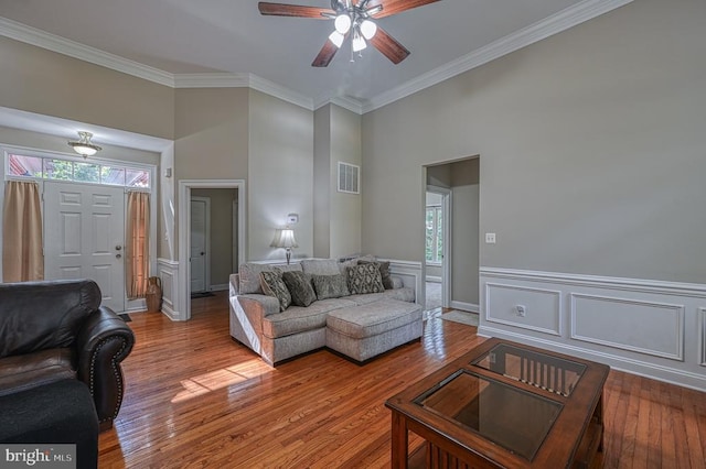 living room with a towering ceiling, light wood-type flooring, ceiling fan, and crown molding