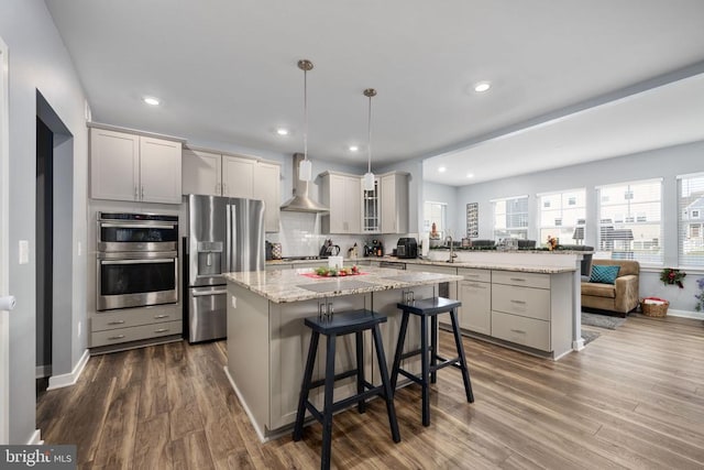 kitchen with a kitchen breakfast bar, wall chimney exhaust hood, stainless steel appliances, pendant lighting, and dark hardwood / wood-style floors