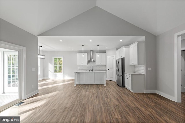 kitchen with pendant lighting, hardwood / wood-style floors, a center island with sink, wall chimney range hood, and stainless steel refrigerator