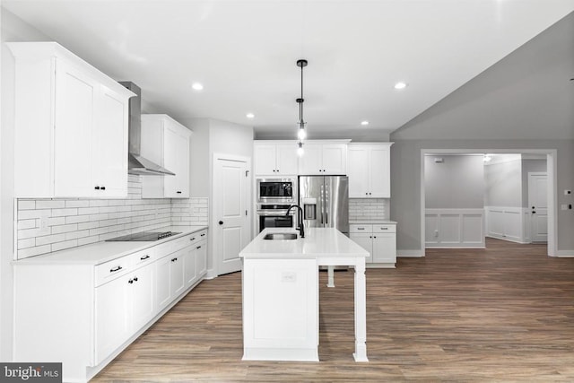 kitchen with wall chimney exhaust hood, stainless steel appliances, decorative light fixtures, white cabinets, and an island with sink