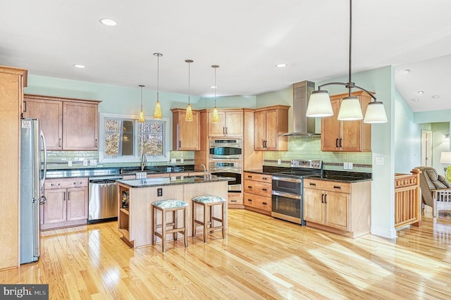 kitchen featuring stainless steel appliances, wall chimney range hood, dark stone countertops, a kitchen bar, and a kitchen island with sink