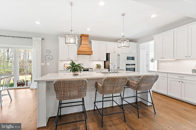 kitchen featuring custom exhaust hood, a center island with sink, white cabinets, hanging light fixtures, and appliances with stainless steel finishes