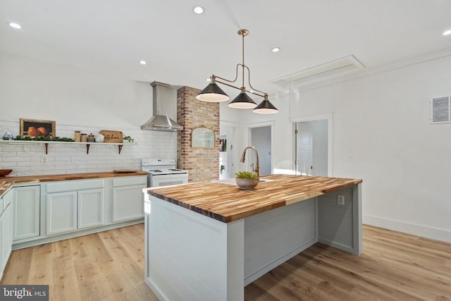 kitchen featuring wall chimney exhaust hood, white electric stove, butcher block countertops, pendant lighting, and a center island with sink