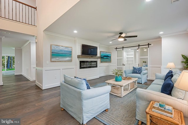 living room with dark wood-type flooring, a barn door, crown molding, a fireplace, and ceiling fan