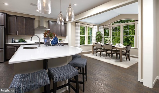 kitchen featuring a center island with sink, decorative light fixtures, dark brown cabinets, wall chimney exhaust hood, and sink