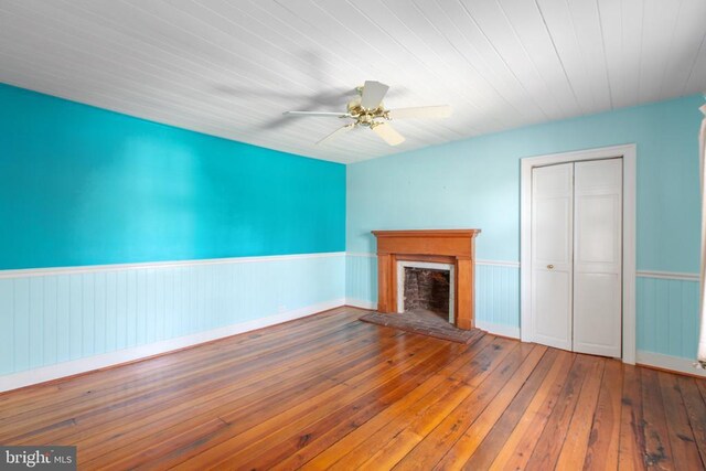 unfurnished living room with wainscoting, a fireplace with raised hearth, ceiling fan, and hardwood / wood-style floors