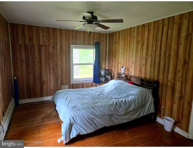 bedroom featuring wood finished floors, wood walls, baseboards, a ceiling fan, and a baseboard heating unit