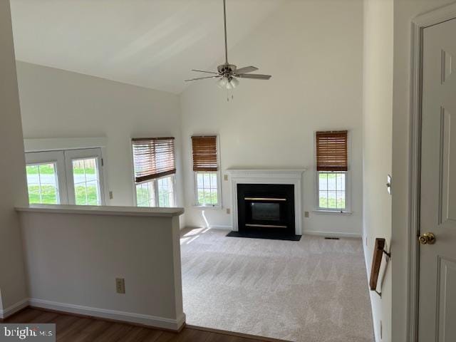 unfurnished living room featuring healthy amount of natural light, ceiling fan, a fireplace with flush hearth, wood finished floors, and high vaulted ceiling