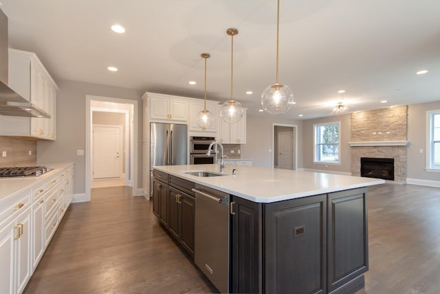 kitchen featuring an island with sink, stainless steel appliances, decorative light fixtures, a stone fireplace, and dark hardwood / wood-style floors