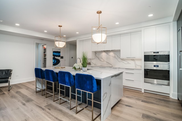 kitchen featuring white cabinets, stainless steel double oven, an island with sink, light wood-type flooring, and pendant lighting