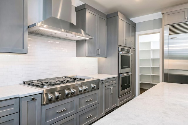 kitchen featuring light stone counters, tasteful backsplash, stainless steel appliances, gray cabinets, and wall chimney exhaust hood