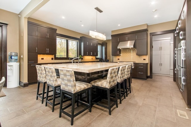 kitchen featuring light tile floors, a center island with sink, backsplash, and dark brown cabinetry