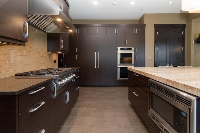 kitchen featuring built in appliances, wall chimney range hood, tasteful backsplash, dark brown cabinets, and dark tile flooring