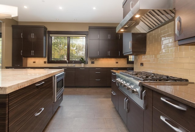 kitchen featuring wall chimney range hood, dark tile flooring, backsplash, stainless steel microwave, and light stone counters