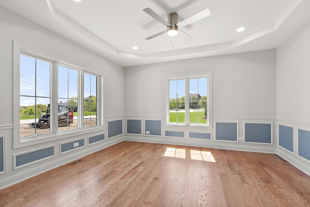 empty room featuring a raised ceiling, healthy amount of natural light, recessed lighting, light wood-style flooring, and a wainscoted wall